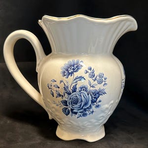 May include: Cream-colored ceramic pitcher with a blue floral design. The pitcher has a curved handle and a scalloped rim with gold trim. The body of the pitcher features a raised floral pattern.