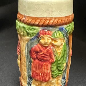 Op de afbeelding: Een cilindrische keramische bierpul met een crèmekleurige boven- en onderkant met een craquelé glazuurafwerking. Het centrale gedeelte toont een reliëfscène van twee figuren in rode en groene kleding, omlijst door boomachtige elementen.