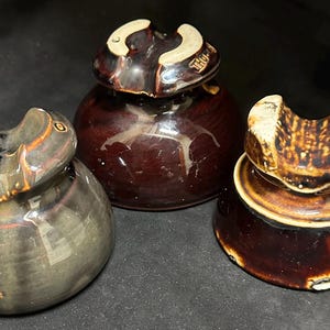 May include: Three antique ceramic insulators in shades of brown and gray. The insulators have a rounded base and a curved top with a groove. The colors range from a dark, glossy brown to a lighter, matte gray.