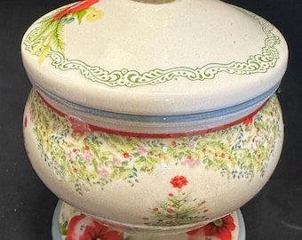 May include: A decorative ceramic jar with a lid and a clear glass knob. The jar is cream-colored with floral patterns in red, green, and yellow. The base and lid have a light blue border.