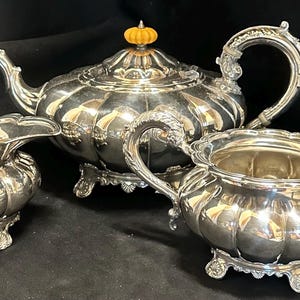Puede incluir: Un juego de té plateado, que incluye una tetera, una jarra de crema y un azucarero, se muestra sobre un fondo oscuro. La tetera tiene un asa decorativa y un pequeño pomo de color naranja en la tapa. El juego tiene un diseño acanalado y asas ornamentadas.