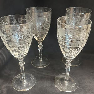 Puede incluir: Cuatro copas de vino de cristal transparente con diseños grabados de flores y hojas. Las copas tienen un cuenco redondeado, un tallo delgado y una base circular. Los patrones grabados incluyen bandas horizontales y motivos botánicos detallados.