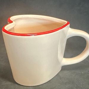 May include: A heart-shaped ceramic mug with a glossy off-white finish and a vibrant red rim along the top edge. The mug features a curved silhouette that forms a heart shape when viewed from above, complete with a sturdy handle on the right side.