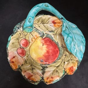 Op de afbeelding: Een keramische serveerschaal met een bladvormig ontwerp, met een handvat en beschilderd met een fruitmotief. De schaal heeft een lichtgroene basis met een grote rode en gele appel, twee kersen en een turquoise handvat en blad.