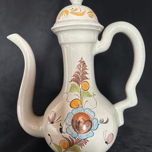 Könnte beinhalten: Cremefarbener Keramikkrug mit Deckel, gebogenem Ausguss und Henkel. Der Krug ist mit handgemalten Blumen- und Schmetterlingsmotiven in Blau-, Braun-, Orange- und Grüntönen verziert. Der Deckel hat ein oranges Wirbeldesign.