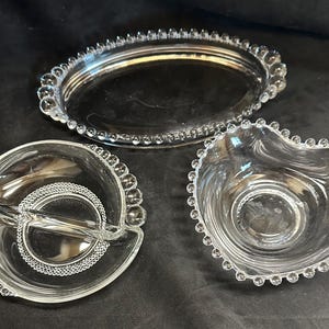 May include: Three clear glass serving dishes. An oval tray with a beaded edge, a heart-shaped bowl with a beaded edge, and a divided dish with a beaded edge and a central circular design.
