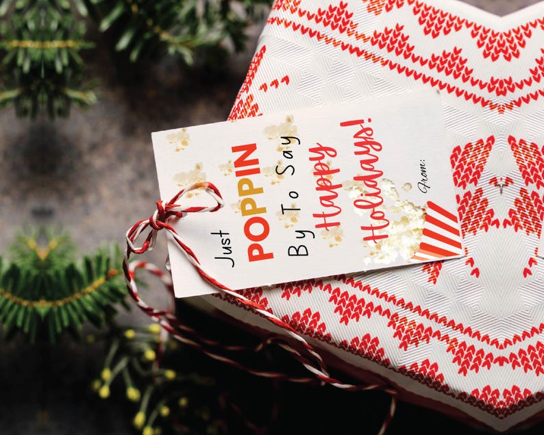 May include: A white gift tag with red and white string tied around it. The tag has gold glitter and the text "Just Poppin' By To Say Happy Holidays!"