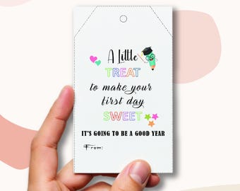 Peut inclure: Une étiquette cadeau blanche avec le texte "A Little TREAT to make your first day SWEET. IT'S GOING TO BE A GOOD YEAR." L'étiquette a une petite illustration d'un diplômé et d'un crayon.