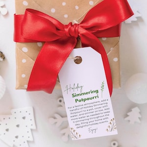 Könnte beinhalten: Ein Geschenkanhänger mit braunem und weißem Polka-Dot-Hintergrund und einem roten Band. Der Anhänger hat ein weißes Etikett mit grünem Text, der "Holiday Simmering Potpourri" lautet. Der Text auf dem Etikett beschreibt, wie man das Potpourri verwendet, um eine festliche Atmosphäre zu schaffen.