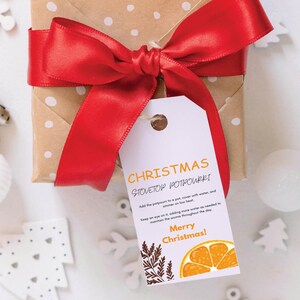 Puede incluir: Una etiqueta de regalo blanca con ilustraciones naranja y marrón y el texto "CHRISTMAS STOVETOP POTPOURRI" e instrucciones de uso. La etiqueta está atada a una caja de regalo marrón con una cinta roja.