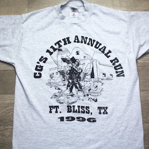 May include: A light gray t-shirt with a black graphic commemorating the "CG's 11th Annual Run" at Ft. Bliss, TX, in 1996. The graphic features a soldier, military vehicles, and a building. The text is in a bold, sans-serif font.