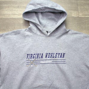 Pode incluir: Camisola cinzenta com capuz com o texto "VIRGINIA WESLEYAN" em azul, com tacos de hóquei cruzados. A camisola tem um capuz com cordão e um bolso canguru. O tecido parece macio e confortável.