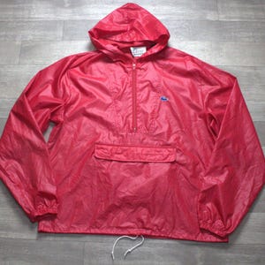 May include: A bright red hooded windbreaker jacket featuring a half-zip front closure, long sleeves with elastic cuffs, and a large front kangaroo pocket. A small blue crocodile logo is embroidered on the chest, and a white label is at the neck.