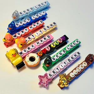 Può includere: Una collezione di mollette decorative in vari colori, ognuna adornata con un ciondolo diverso e una frase motivazionale scritta con perline di lettere bianche. Le frasi includono "GOOD LUCK" e "WORK HARD".