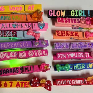 Pode incluir: Uma coleção de molas de roupa decorativas de madeira em várias cores, incluindo amarelo, laranja, rosa, roxo e azul. Cada mola apresenta texto como "OMG LOOK AT" e "GLOW GIRL!" juntamente com enfeites como laços e berloques.