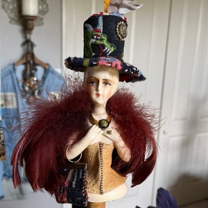 May include: A vintage doll with a painted face and a colorful hat with feathers. The doll is wearing a brown corset and a red feather boa. The doll is standing on a green pedestal.