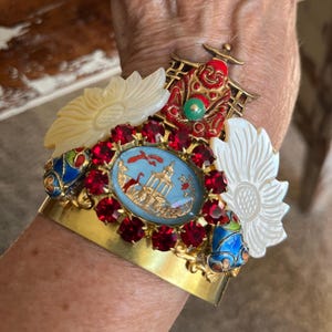 May include: A gold-toned cuff bracelet with ornate details. It features a central oval design with a light blue background and a gold building illustration, surrounded by red gemstones. Additional embellishments include white flower-shaped accents and colorful enamel details.