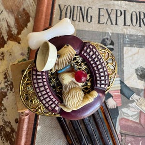 May include: A decorative hair comb featuring a sculpted beige dog figure with a blue collar and a red bead charm. The dog is positioned within a dark purple circular frame, accented by a white bone, a small white bowl, and gold-tone filigree.