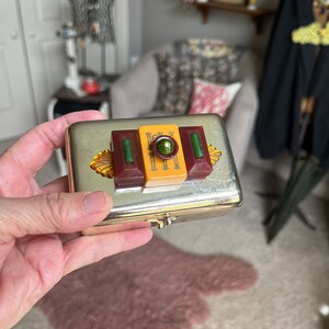 Puede incluir: Una caja de metal plateado con una tapa con bisagras. La caja tiene un dise&ntilde;o decorativo en la parte superior con tres piezas rectangulares de pl&aacute;stico en rojo, amarillo y blanco. Las piezas de pl&aacute;stico tienen un dise&ntilde;o verde y dorado.