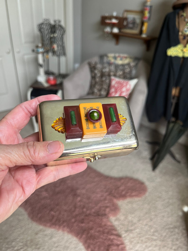 Puede incluir: Una caja de metal plateado con una tapa con bisagras. La caja tiene un dise&ntilde;o decorativo en la parte superior con tres piezas rectangulares de pl&aacute;stico en rojo, amarillo y blanco. Las piezas de pl&aacute;stico tienen un dise&ntilde;o verde y dorado.