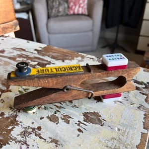 May include: A vintage wooden clothespin with a yellow sign that reads "AGRICULTURE". The clothespin has a metal spring and a small, white and red block on top. The clothespin is on a distressed, white-painted wooden surface.