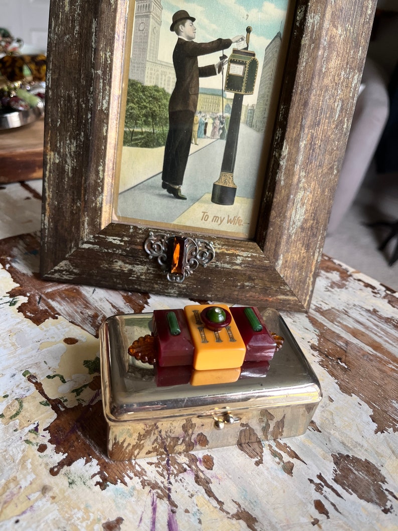 Puede incluir: Una caja de metal plateado con una tapa con bisagras. La caja tiene un dise&ntilde;o decorativo en la tapa hecho de pl&aacute;stico amarillo, rojo y verde. La caja est&aacute; sobre una superficie de madera.