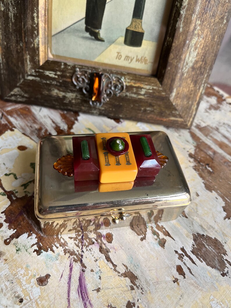 Puede incluir: Una caja de metal plateado con una tapa con bisagras. La tapa est&aacute; decorada con tres capas de pl&aacute;stico de colores, incluyendo rojo, amarillo y verde. La caja tiene un peque&ntilde;o pomo dorado en la parte delantera.