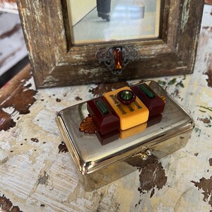 Puede incluir: Una caja de metal plateado con una tapa con bisagras. La caja est&aacute; decorada con un mosaico colorido de piezas de pl&aacute;stico en tonos rojos, amarillos y verdes.