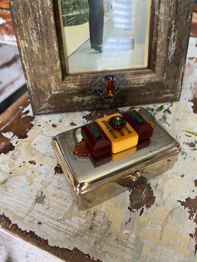 Puede incluir: Una caja de metal plateado con una tapa con bisagras. La caja est&aacute; decorada con un mosaico colorido de piezas de pl&aacute;stico en tonos rojos, amarillos y verdes.