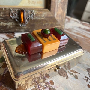 Puede incluir: Una caja de metal plateado con una tapa con bisagras. La caja est&aacute; decorada con un bot&oacute;n de estilo vintage hecho de pl&aacute;stico rojo, amarillo y verde. El bot&oacute;n tiene un centro verde con un peque&ntilde;o strass transparente.