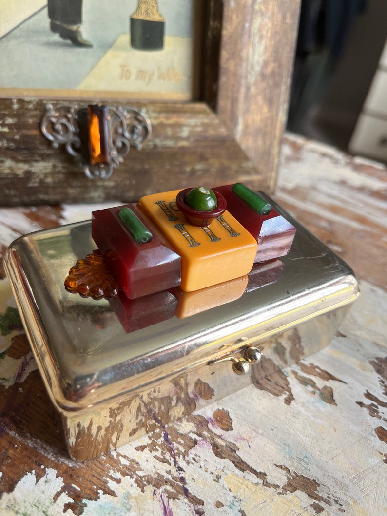 Puede incluir: Una caja de metal plateado con una tapa con bisagras. La caja est&aacute; decorada con un bot&oacute;n de estilo vintage hecho de pl&aacute;stico rojo, amarillo y verde. El bot&oacute;n tiene un centro verde con un peque&ntilde;o strass transparente.