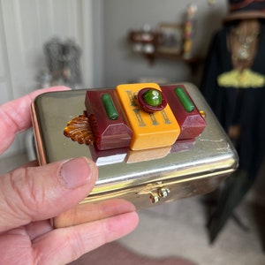 Puede incluir: Una caja de metal plateado con una tapa con bisagras. La tapa est&aacute; decorada con un dise&ntilde;o geom&eacute;trico colorido hecho de pl&aacute;stico rojo, amarillo, verde y &aacute;mbar. El dise&ntilde;o presenta un c&iacute;rculo verde central con un patr&oacute;n en relieve.