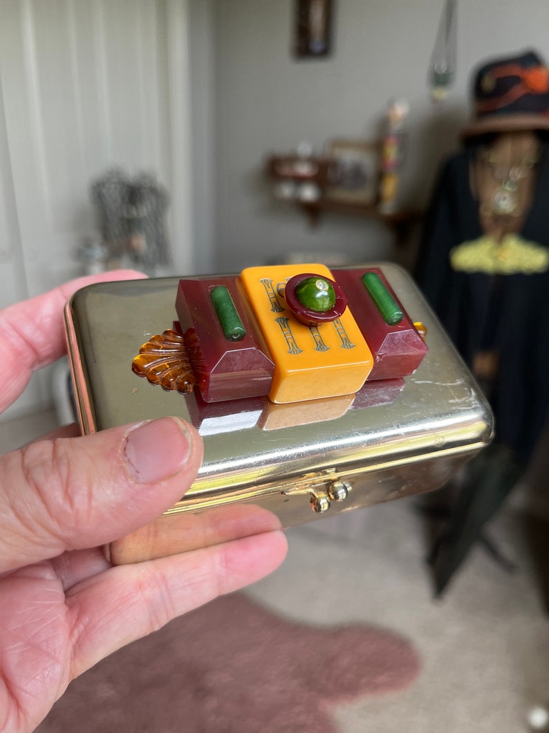 Puede incluir: Una caja de metal plateado con una tapa con bisagras. La tapa est&aacute; decorada con un dise&ntilde;o geom&eacute;trico colorido hecho de pl&aacute;stico rojo, amarillo, verde y &aacute;mbar. El dise&ntilde;o presenta un c&iacute;rculo verde central con un patr&oacute;n en relieve.