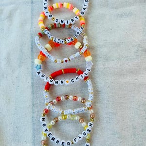 May include: A stack of colorful beaded bracelets with words like "Plaid Shirt Days", "The Sky Is Gold", "Autumn Chill", "November Flush", and "Flannel Cure".
