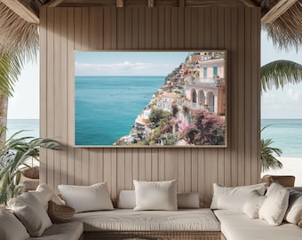 Pode incluir: Uma impressão emoldurada de uma cidade costeira em um penhasco com vista para o mar turquesa. A impressão é exibida em uma parede de madeira dentro de uma área de estar externa coberta. A cena inclui edifícios, barcos e vegetação exuberante.