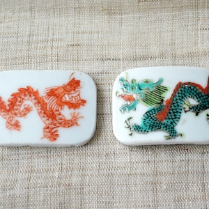 May include: Two rectangular white ceramic tiles with dragon designs. One tile features a red dragon, the other a teal dragon with red accents. The dragons are detailed and stylized, with visible scales and features.