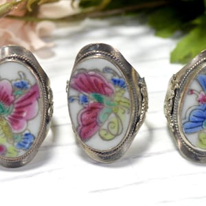 May include: Three oval rings featuring white enamel centers with colorful butterfly designs in pink, blue, and green. Each ring has a silver-toned metal band with decorative twisted wire detailing around the oval setting.