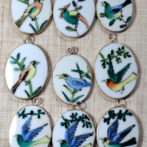 May include: Nine oval-shaped pendants featuring hand-painted bird illustrations on a white background. Each pendant is framed in a metallic border and includes a small silver-toned jump ring at the top for attachment to a necklace or chain.