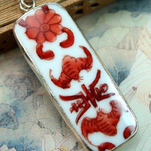 Könnte beinhalten: Ein weißer Porzellananhänger mit silbernem Rand, verziert mit roten, gemalten Motiven einer Blume, zwei stilisierten Tieren und chinesischen Schriftzeichen.