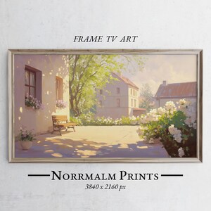 May include: A painting of a sunny courtyard with a white building, a bench, and a flower bush. The building has a window with a flower box and a door. The courtyard is paved with stone and has a green lawn. The painting is framed in a light wood frame.