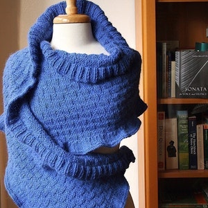 May include: A blue knitted shawl with a cowl neck. The shawl is draped over a mannequin and is displayed against a bookshelf.
