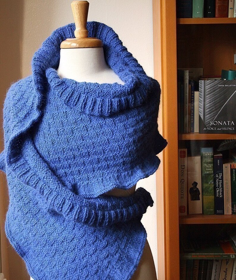 May include: A blue knitted shawl with a cowl neck. The shawl is draped over a mannequin and is displayed against a bookshelf.