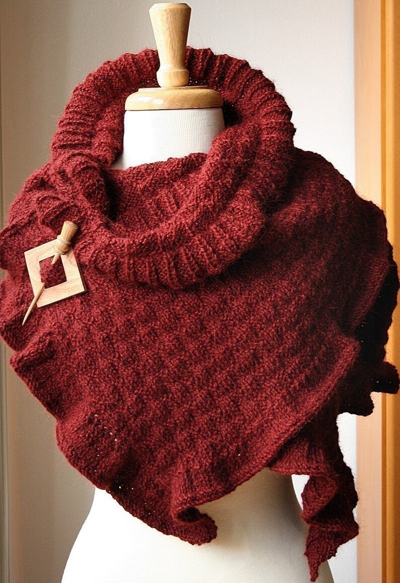 May include: A maroon knitted cowl scarf with a wooden pin and a decorative wooden piece. The scarf is draped over a white mannequin.