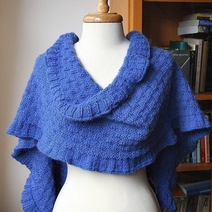 May include: A blue knitted shawl with a ruffled edge, draped over a white mannequin. The shawl is made with a textured stitch pattern.