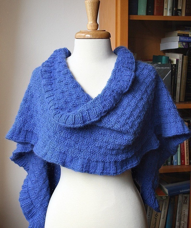 May include: A blue knitted shawl with a ruffled edge, draped over a white mannequin. The shawl is made with a textured stitch pattern.