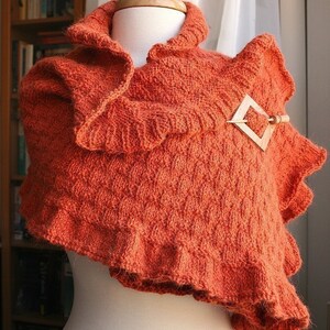 May include: A burnt orange knitted shawl with a ruffled edge and a wooden pin. The shawl is draped over a white mannequin.