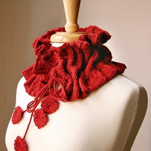May include: A red crocheted cowl with a ruffled edge and a decorative flower accent. The cowl is displayed on a white mannequin.
