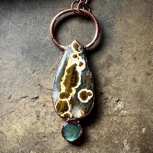 May include: A handcrafted copper pendant necklace. The pendant features a teardrop-shaped stone with brown, white, and gray patterns, and a small, round, teal-colored stone at the bottom. The necklace has a copper chain and a large copper ring.