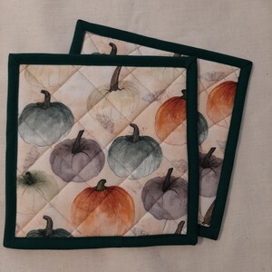 May include: Two quilted pot holders with a dark green border. The pot holders feature a pattern of watercolor pumpkins in shades of orange, green, and gray on a cream background.