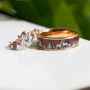 May include: A set of two wedding bands. The engagement ring features a large, clear, oval-shaped diamond set in a delicate, leafy band. The wedding band is a wide, brown band with a deer silhouette design.